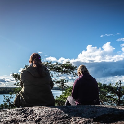 Kaksi naista istuu korkealla kalliolla ja katselee kohti merta maisemaa.