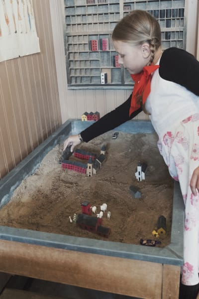Flicka leker bygger liten by av träklossar i sandlåda.