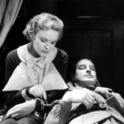 Vaaleahiuksinen nainen on kytketty käsiraudoilla nukkuvaan tai tajuttomaan mieheen: nainen on tiirikoimassa rautoja auki, kuva Hitchcockin elokuvasta 39 askelta.