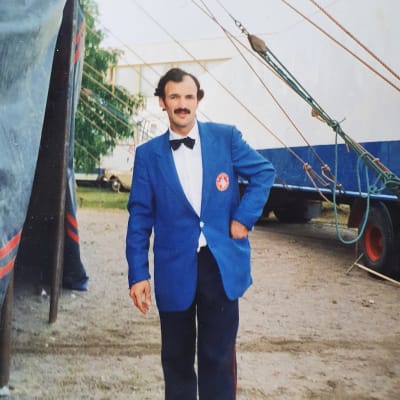 Viiksekäs mies seisoo vasenta kättä puvuntakin taskuun työntäen sirkuksen pihalla.