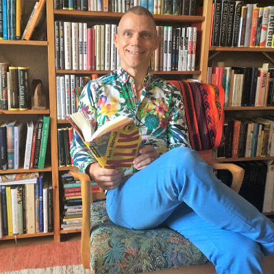 Jari Järvelä istuu tuolissa kirjahyllyn edessä kirja kädessä 