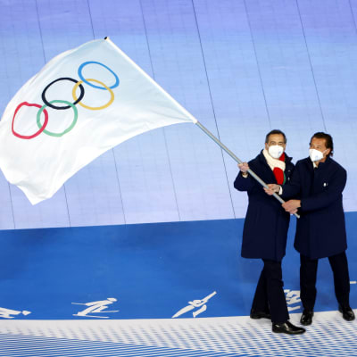Kaksi miestä pitelee suurta olympialippua, kolmas katselee vierestä.
