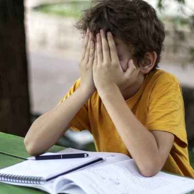 Ett barn sitter vid ett bord med skolböcker framför sig. Barnet håller sitt ansikte i sina händer och ser ledset ut.