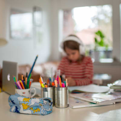 Ett barn sitter vid en dator med hörlurar på huvudet. Framför på bordet ligger skolböcker och burkar fyllda med olika sorters pennor.