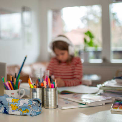 Ett barn sitter vid en dator med hörlurar på huvudet. Framför på bordet ligger skolböcker och burkar fyllda med olika sorters pennor.