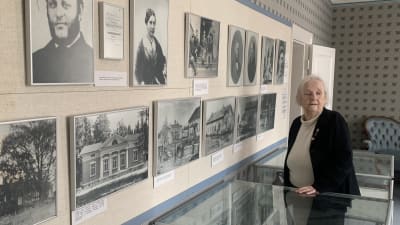 En äldre kvinna står i ett musem vid en vägg fullt av gamla fotografier. 