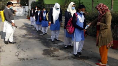 Elever i skoluniform stör i kö för att få temperaturen kontrollerad av en kvinna i brun yllejacka. En man med en gul behållare på ryggen sprutar desinfektionsmedel på marken runt elevernas fötter.
