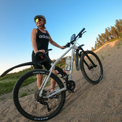 Anneli Magnusson står på en grusväg och håller i en stående terrängcykel. Hon har på sig träningskläder, cykelhjälm och solglasögon