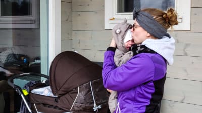 En kvinna i lila jacka håller en baby i famnen. Hon ser ut att pussa babyns hjässa eller tala lugnande till babyn som ser lite missnöjd ut.