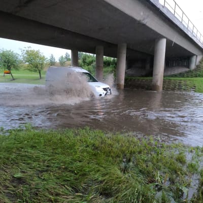 En vit minivan kör genom djupt flödvatten under en bro. 