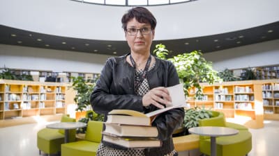 Minna Koistinen, en dam med glasögon och svart läderjacka, håller i en hög med böcker i ett bibliotek.
