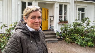 En kvinna i svart jacka står framför ett vitt hus och tittar in i kameran.