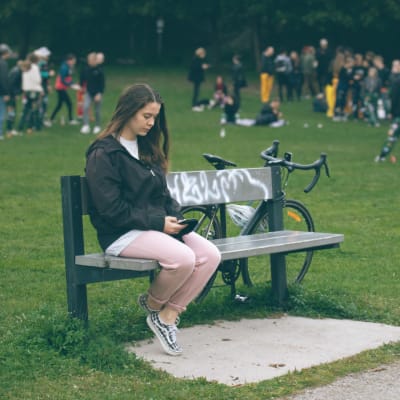 Nuori nainen istuu yksin puistonpenkillä älypuhelin kädessään. Taustalla muut nuoret pitävät hauskaa