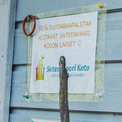 Ljusblå husvägg med skylt där det står att trädgårdsodlingen handhas av daghemsgruppen Sateenkaari Koto.
