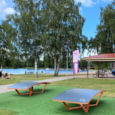Lähikuvassa on Heinolan uimaranta ja rantapuisto. Ihmisiä istumassa nurmikolla ja kuvassa myös urheiluvälineitä.