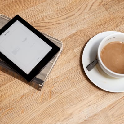 En läsplatta ligger på en bok bredvid en kopp kaffe.