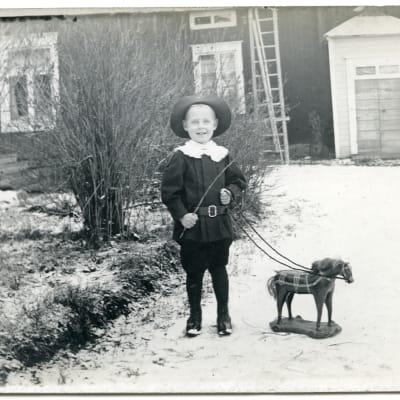En pojke i finkläder leker med en trähäst i tömmar och med vidja som piska. På gårdsplan. 1880-1920-tal.
