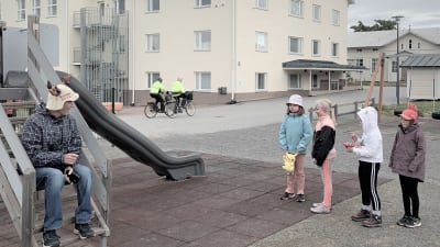 En man med bastuhatt sitter på trappan till en lekställning medan fyra barn står intill på en skolgård. 