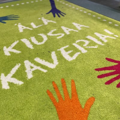 Matto koulun käytävällä, matossa värikkäitä käsiä ja teksti: Älä kiusaa kaveria!