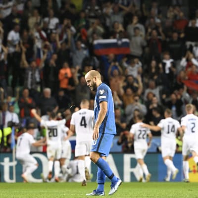 Teemu Pukki tittar ner i marken medan slovenska spelare firar i bakgrunden.