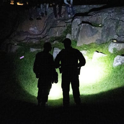 Kaksi poliisia osoittaa taskulampuilla pimeyteen Helsingin Kaivopuistossa. Poliisit näkyvät siluetteina, taustalla nurmea ja kalliota.