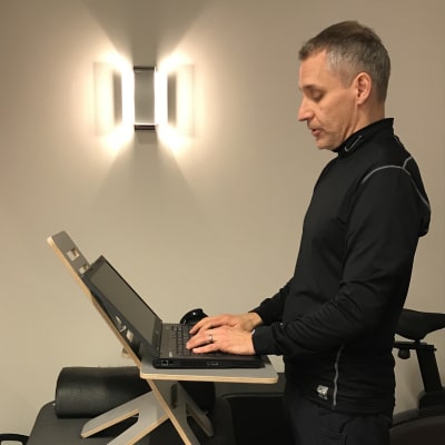 En man står vid sin laptop som ligger på ett litet bord. Bordet i sin tur står på en bänk så mannen får en optimal ståend arbetsställning.
