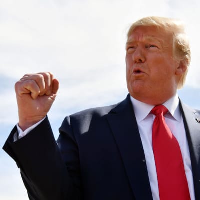 Donald Trump reagerar genom att knyta bägge händer. Han är fotad utomhus, iklädd mörkblå kostym, vit skjorta och röd slips. 