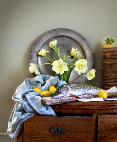 Fotografi av ett stilleben med en gädda, salt, citroner och blommor i vas på en byrå.
