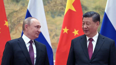 Två äldre män i mörk kostym och violett slips tittar snett på varandra. I bakgrunden Kinas och Rysslands flaggor.