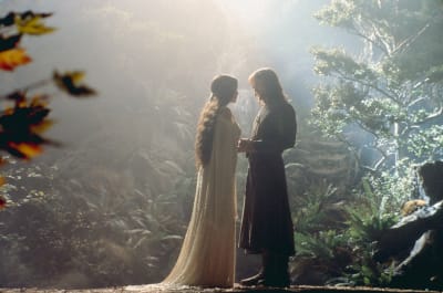 En kvinna i klänning och en man i lång rock står i ett skogslandskap. 