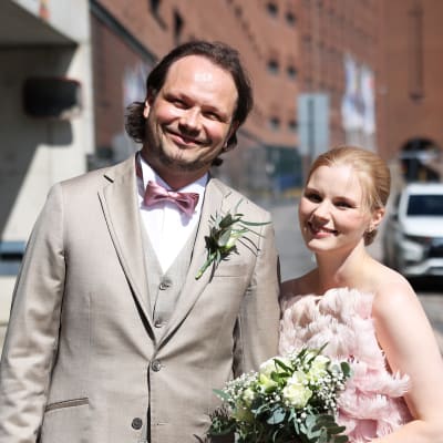 Tuleva aviopari hymyilee ja katsoo kameraan juhlavaatteet päällään maistraatin edessä Helsingissä. Taustalla lintulahdenkuja.