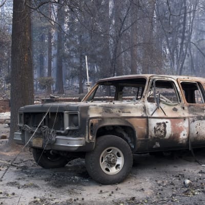 I förgrunden en bil som förstörts i skogsbrand, i bakgrunden ruinerna efter en byggnad.