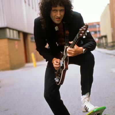 Brian May från Queen poserar 1989 på en skateboard med gitarr i handen ute på en gata.