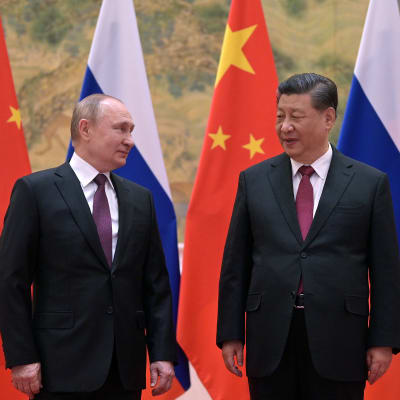 Vladimir Putin och Xi Jinping står bredvid varandra och tittar på varandra. I bakgrunden flaggor.