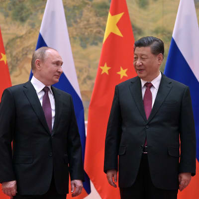 Vladimir Putin och Xi Jinping står bredvid varandra och tittar på varandra. I bakgrunden flaggor.