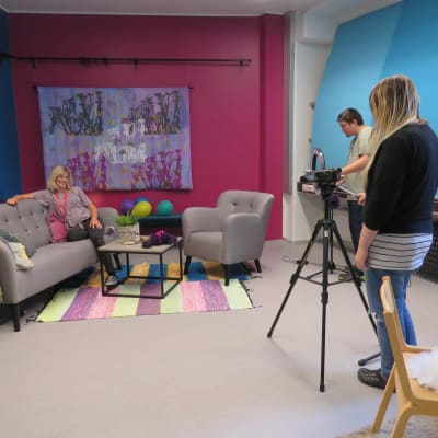 Kårkullas tv-studio. Två personer filmar en tredje person som sitter i en soffa i ett färggrant rum.