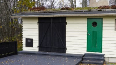 En stransbastu med ljusbeige fasad, svarta skjutdörrar och en grön dörr. Bastun har grästak. På fasaden finns en solfångare monterad.