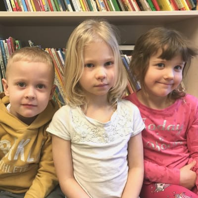 Tre barn i dagisåldern framför en bokhylla med barnböcker.