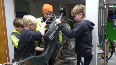 Fyra pojkar i ålder 10-12 skruvar isär en gammal moped. De är inne i en verkstad.