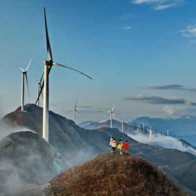 En grupp människor står på en topp i en bergskedja som bekläs av vindturbiner. 