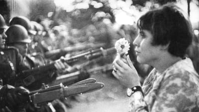 En hippiekvinna står med en blomma i handen inför beväpnade män.