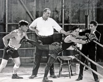 Buster Keaton nyrkkeilijänä roikkuu kehän köysissä ja pitelee vastustajaa kauempana toisella jalallaan tuomarin ihmetellessä. Kuva elokuvasta Rakkaus ja romahdus