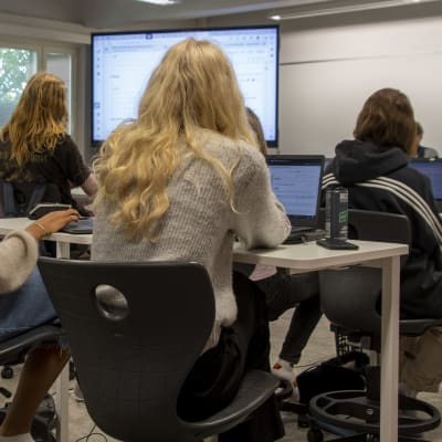 Några personer sitter i ett klassrum med sina datorer på borden. De tittar mot en stor skärm längst fram i klassen.