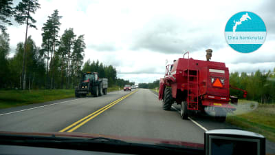 En tröska och en traktor stoppar upp trafiken i båda filerna på den asfalterade vägen som omges av skog. Uppe i högra hörnet finns en stämpel där det står Dina hemknutar.