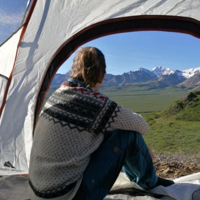 Nainen katsoo teltan oviaukosta ulos, taustalla vuoret