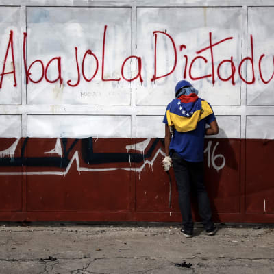 En demonstrant håller i stenar i sammandrabbning med polis i Venezuela. På väggen står "Ner med diktaturen" på spanska.  