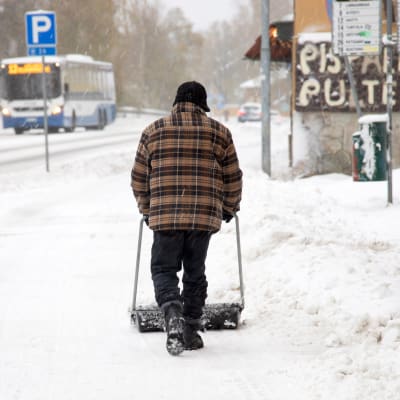 Henkilö kolaa lunta jalkakäytävällä  lumisessa maisemassa.