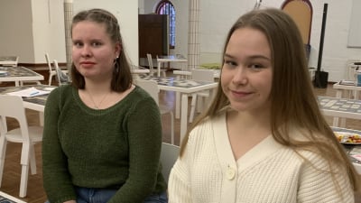 Två unga kvinnor med långt hår sitter på var sin stol i ett stort rum. 