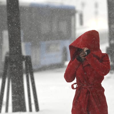 En rödklädd person med huvan uppdragen över ansiktet vandrar förbi en buss i snöyra.