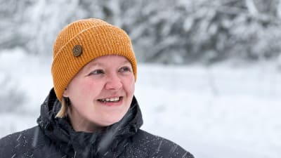 En dam med gul mössa står i snöfallet och tittar leende åt sidan.